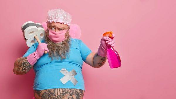 Verspielter Mann in Reinigungsoutfit mit Schrubber und Sprühflasche bereit für die Hausreinigung