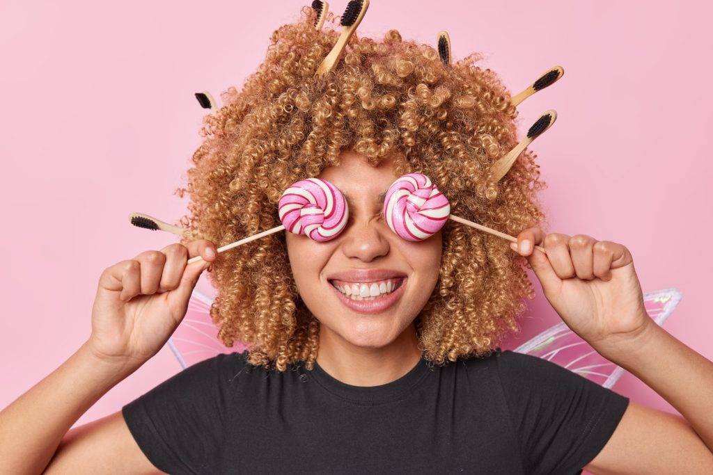 Fröhliche junge Frau mit Lollipops vor den Augen, umgeben von Make-up Pinseln, symbolisiert spielerische Seniorenunterstützung.